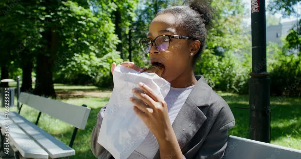 A woman in formal clothes eats a croissant and drinks coffee from a disposable cup while sitting on a bench in a city park. A sunny summer day A woman in formal clothes eats a croissant and drinks coffee from a disposable cup while sitting on a bench in a city park. A sunny summer day