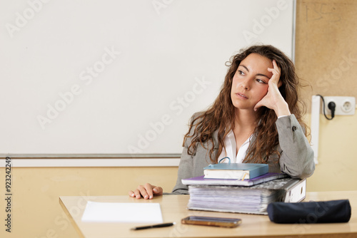 Woman overwhelmed by work overload at the table in her office or high school classroom, she looks to the side thinking where to start.