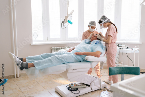 Wide shot of female surgeons carry out hair FUE Sapphire method hair transplant surgery in operating room to male patient with hair loss problem. Concept of professional baldness treatment. photo