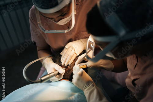 Close-up of two female doctors in lighting glasses extracting hair follicles from donor area of male patient for hair transplantation in clinic. Process of hair transplant with FUE Sapphire method.