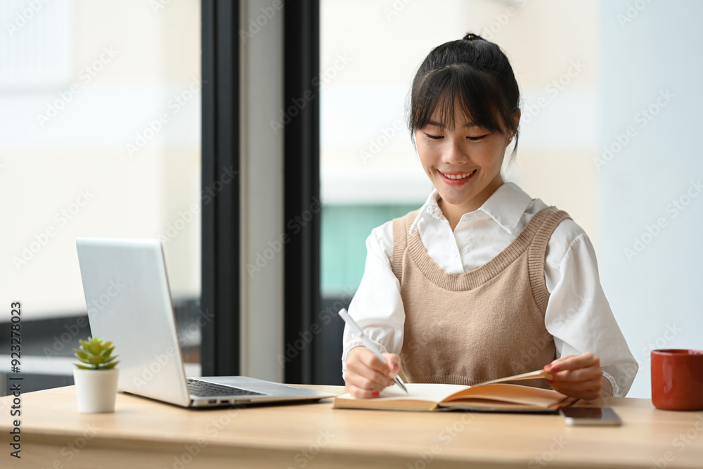 Smiling asian female employee working on laptop, making notes in cozy workspace Smiling asian female employee working on laptop, making notes in cozy workspace