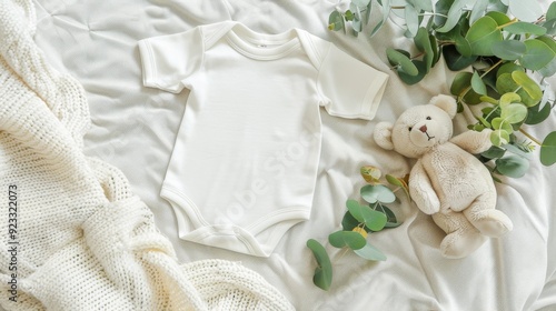 A soft, neutral-colored baby onesie lies on a bed of blankets next to a plush teddy bear and eucalyptus branches, creating a serene and cozy baby scene.