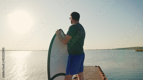 A man standing on a lake pier at sunset, leaning on a water board. Male surfer holding a short surfboard while standing on a pier with a motorboat in the background photo