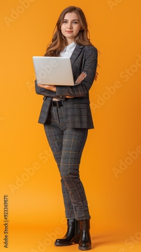 A confident young woman stands with her arms crossed, holding a laptop, dressed in a tailored suit