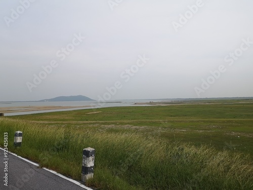 A scenic summer landscape featuring a winding road through a green field with mountains, a river, and a coastal horizon under a cloudy sky