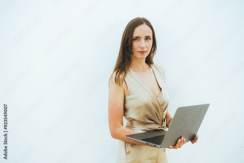 A woman standing against a light-colored wall, holding an open laptop with both hands, dressed in a beige sleeveless dress, looking directly at the camera A woman standing against a light-colored wall, holding an open laptop with both hands, dressed in a beige sleeveless dress, looking directly at the camera