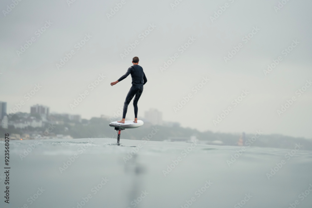 A young man in a wetsuit rides in the sea above the water on an electric powered hydrofoil board, efoil, e-foil, foil drive, hybrid foil. A young man in a wetsuit rides in the sea above the water on an electric powered hydrofoil board, efoil, e-foil, foil drive, hybrid foil.