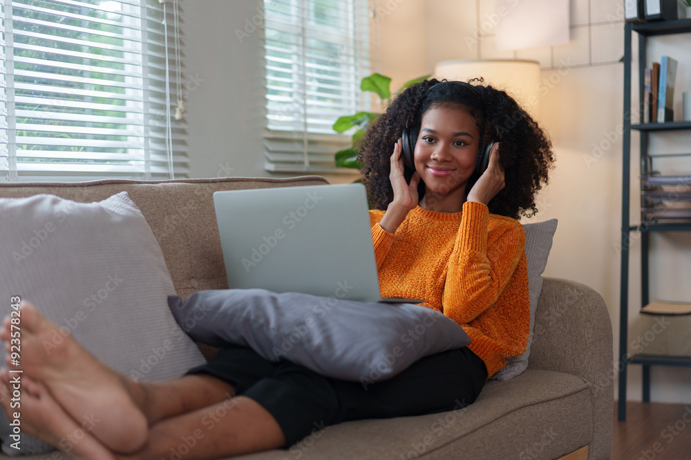 Smiling African American woman relaxing on a cozy couch, wearing headphones, using a laptop, enjoying a comfortable home. Smiling African American woman relaxing on a cozy couch, wearing headphones, using a laptop, enjoying a comfortable home.