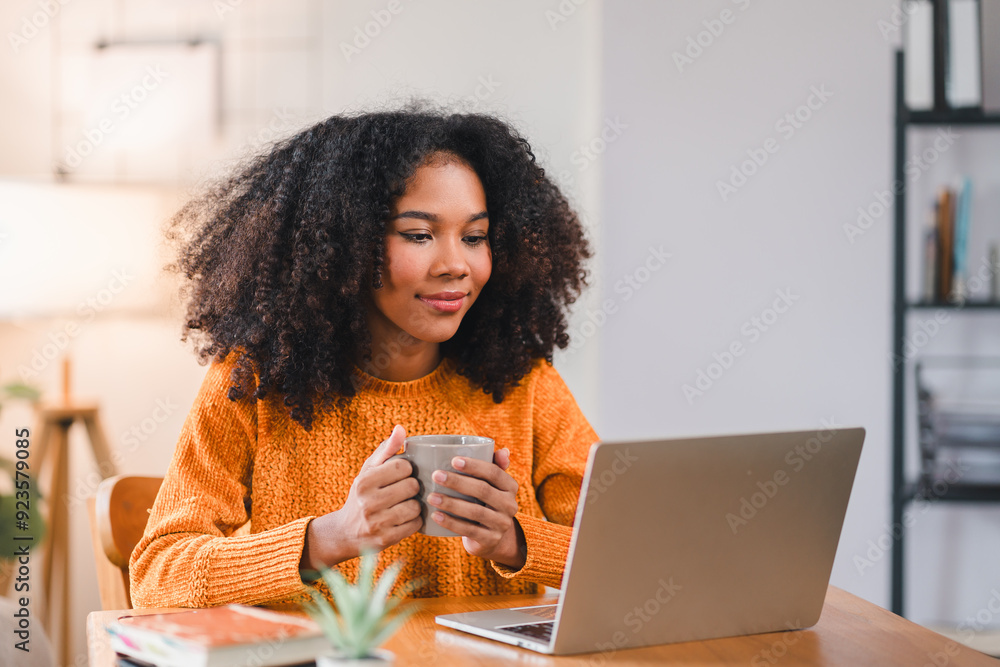 Focused African American woman holding a coffee mug while working on a laptop at home, sitting at a desk with books. Focused African American woman holding a coffee mug while working on a laptop at home, sitting at a desk with books.