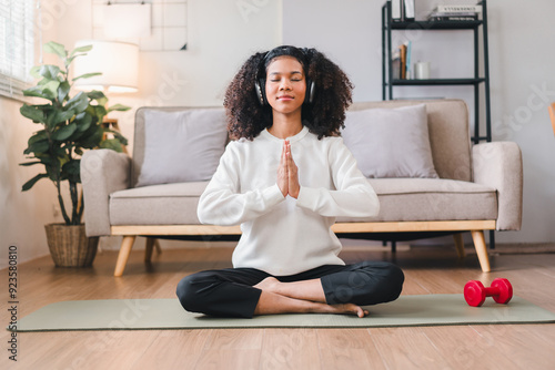 Relaxed African American woman meditating in seated position on yoga mat at home, listening to music with headphones.