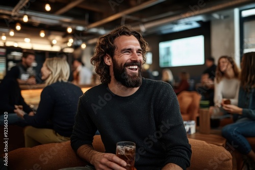 A man happily laughs while holding a drink in a lively indoor setting, surrounded by friends and filled with warm lights, reflecting joy and camaraderie.