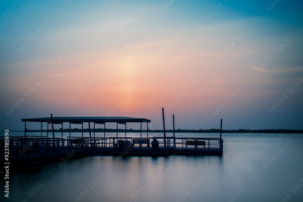 Obraz premium A serene sunset over a tranquil Lewisville lake in The Colony, TX, with a silhouetted pier reflecting soft hues of blue, pink, and orange, creating a peaceful, calming atmosphere