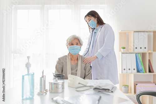 Female doctor in white coat comforting elderly patient in bright medical office. Both wearing face masks. doctor use stethoscope listening lung of patient, and pandemic safety measures in hospital.