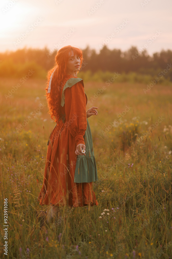 Fototapeta premium A shepherdess woman with red hair and a red and green dress walks on a field of flowers. Rural scene concept with a romantic peasant woman in a linen dress. Romance novel.