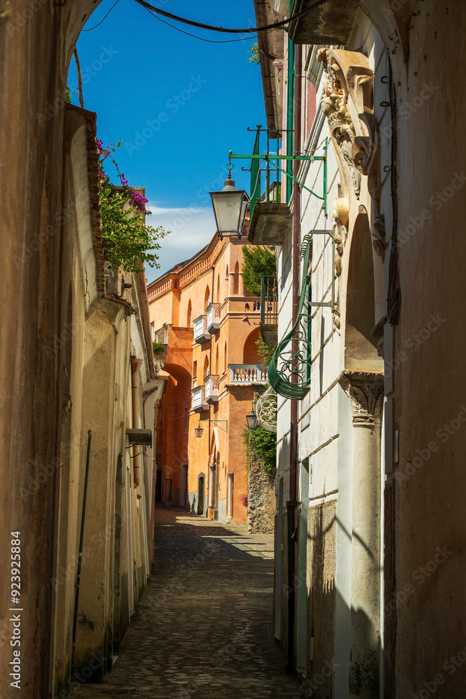 A picturesque narrow alley in a charming Italian village, bathed in warm sunlight. The vibrant orange and terracotta facades of the buildings, adorned with ornate architectural details,