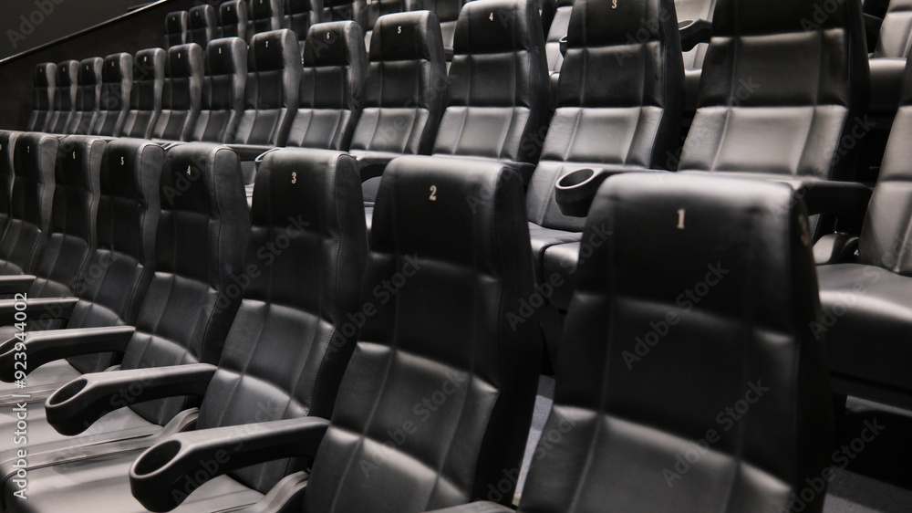 Naklejka premium Rows of empty black leather cinema seats with cup holders and numbered backrests in a modern movie theater.