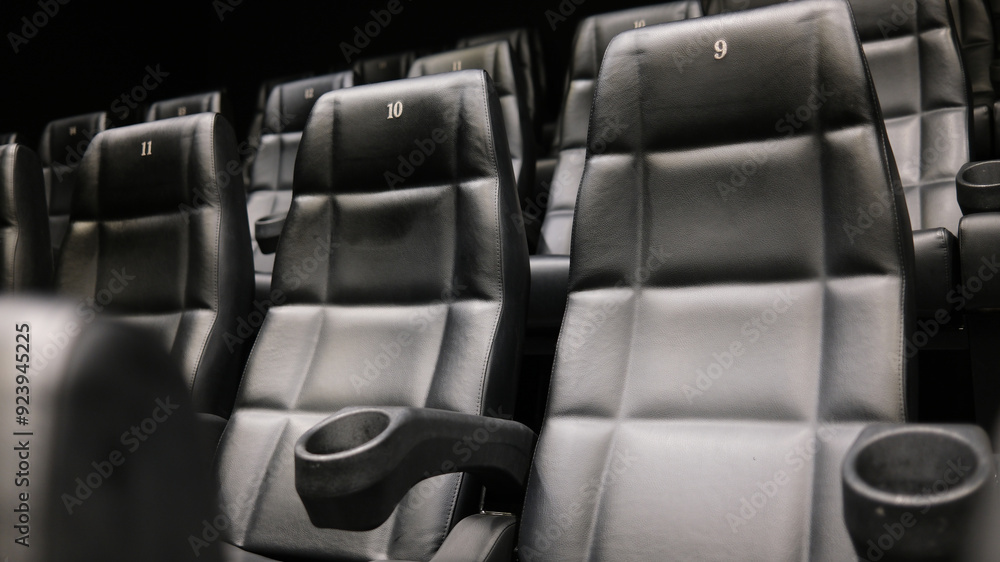 Rows of empty black leather cinema seats with cup holders and numbered backrests in a modern movie theater. Rows of empty black leather cinema seats with cup holders and numbered backrests in a modern movie theater.
