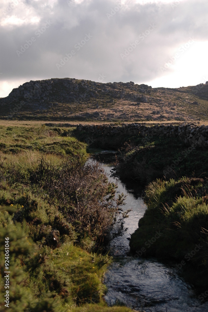 A tranquil stream flows through a rugged landscape, with green vegetation lining the banks and a rocky hill A tranquil stream flows through a rugged landscape, with green vegetation lining the banks and a rocky hill
