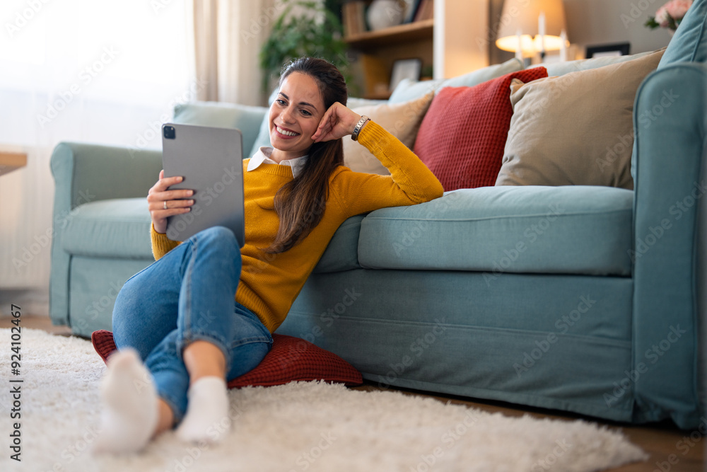 Obraz premium Relaxed, cheerful woman uses a tablet for a video call while sipping coffee, cozily seated on the floor by the couch in her living room.