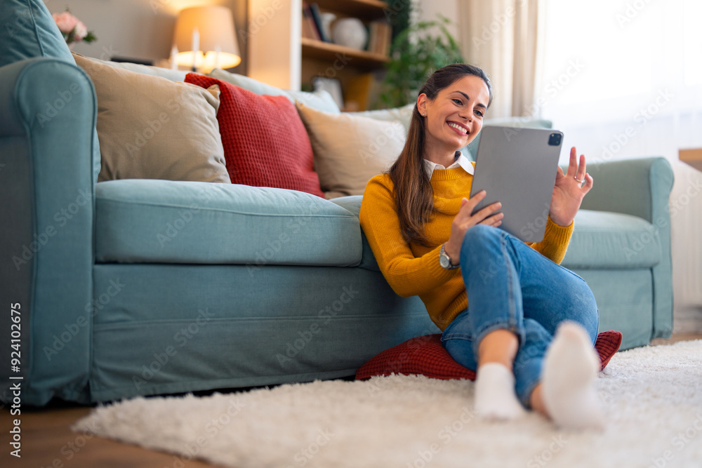 Obraz premium A cheerful caucasian woman enjoys a video chat on her tablet, sipping coffee and sitting casually on the floor by the couch in her cozy living room.