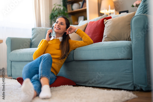 A joyful Caucasian woman engages in a conversation over the phone, seated on the floor and leaning against a sofa in a cozy living room, dressed in casual attire.