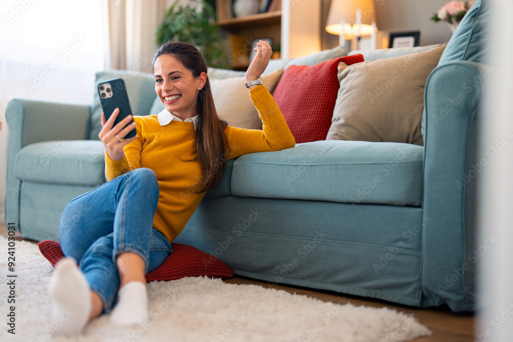 Obraz premium Caucasian woman in casual attire engages in a lively videocall, sitting comfortably on the floor by the couch within a well-lit living room.