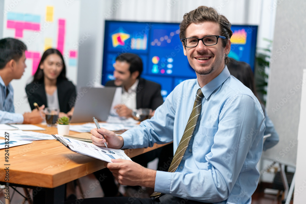 Portrait of businessman or analyst looking at camera with his colleague analyzing data analysis in dynamic business strategy investment planning meeting. Meticulous Portrait of businessman or analyst looking at camera with his colleague analyzing data analysis in dynamic business strategy investment planning meeting. Meticulous