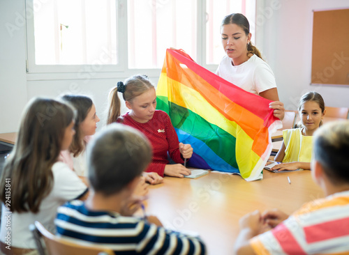 Young progressive female teacher discussing with preteen children about LGBT social movements in classroom, holding rainbow flag photo