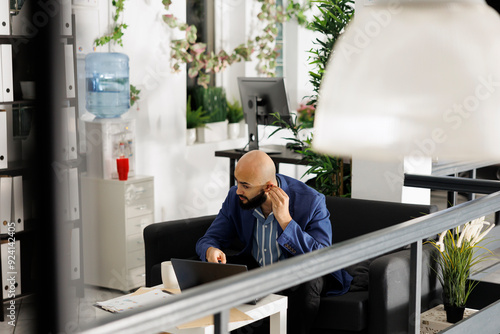 Arab corporate employee putting in earbuds to answer ceo videocall on laptop while working in startup business office room. Professional wearing earphones to talking online in green open space