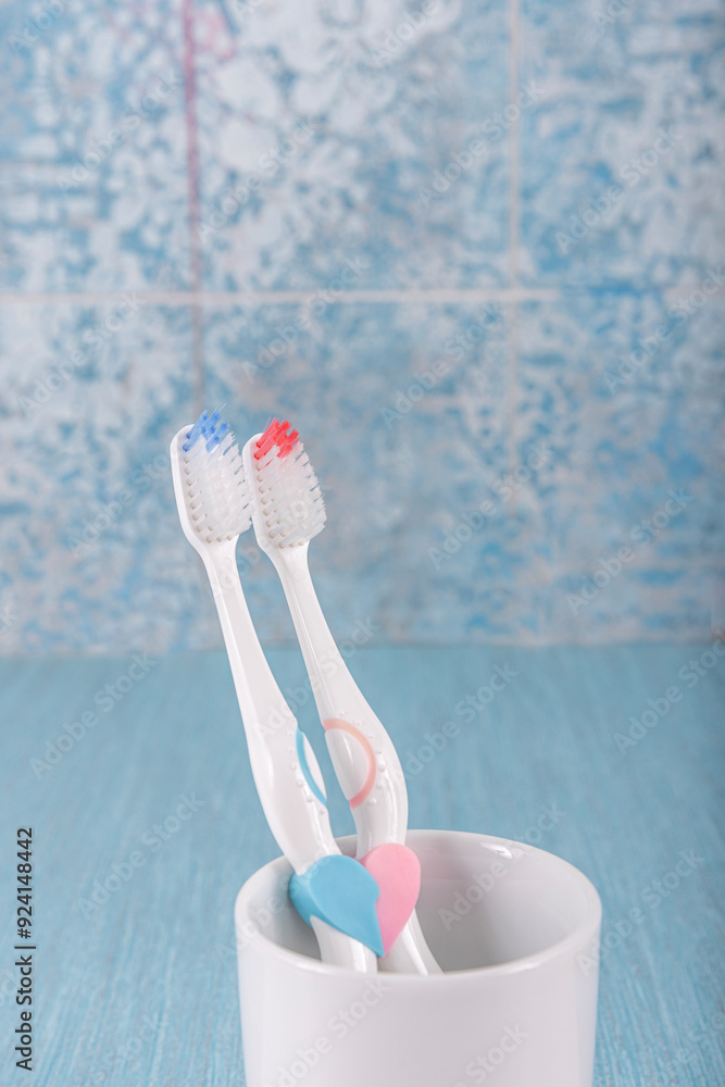 There is a heart pattern on the toothbrush handle. Two toothbrushes in ceramic mug on blue background. Toothbrush holder with toothbrushes, dental hygiene concept. There is a heart pattern on the toothbrush handle. Two toothbrushes in ceramic mug on blue background. Toothbrush holder with toothbrushes, dental hygiene concept.