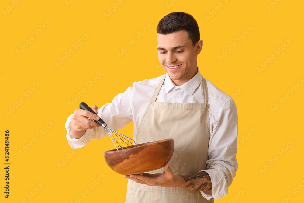 Handsome happy male chef with bowl and whisk on yellow background