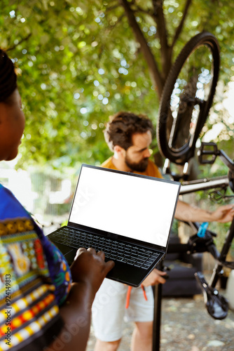 Young african american female using laptop with isolated screen doing bike maintenance at home yard. Minicomputer displaying blank screen while two active sportloving persons repairs broken bicycle. photo