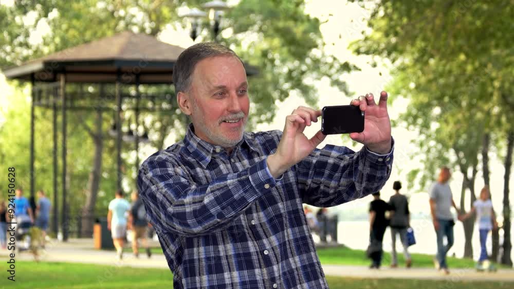 Senior caucasian man taking photo by his smartphone in the park. Sightseeing and photography concept. Senior caucasian man taking photo by his smartphone in the park. Sightseeing and photography concept.