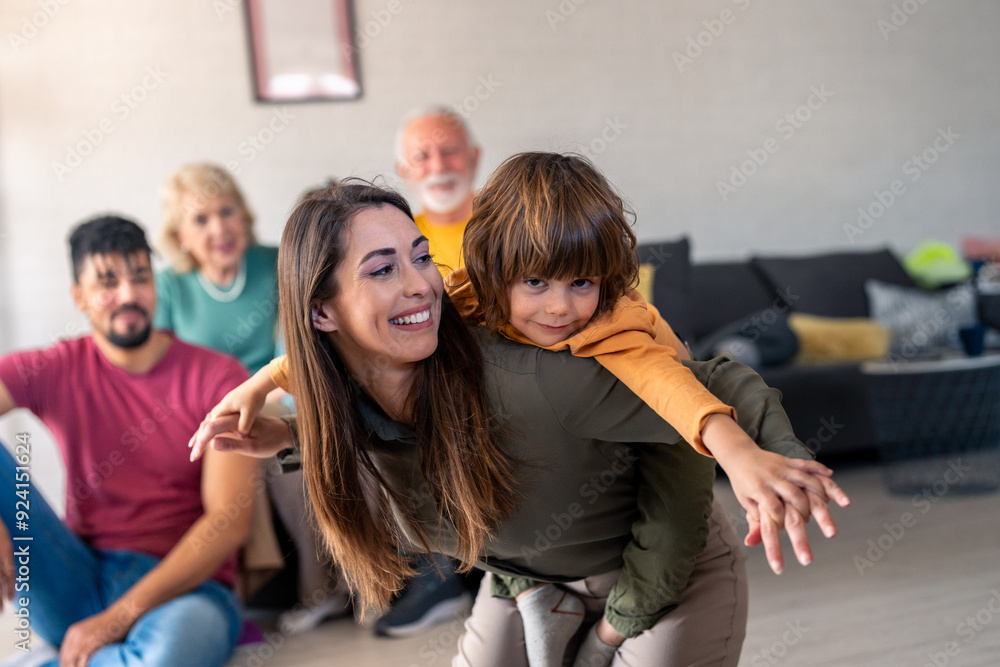 A cheerful woman enjoys piggybacking her young son, while a multi-generational, diverse family relaxes together, sitting and smiling in the cozy ambiance of a well-lit living room. A cheerful woman enjoys piggybacking her young son, while a multi-generational, diverse family relaxes together, sitting and smiling in the cozy ambiance of a well-lit living room.