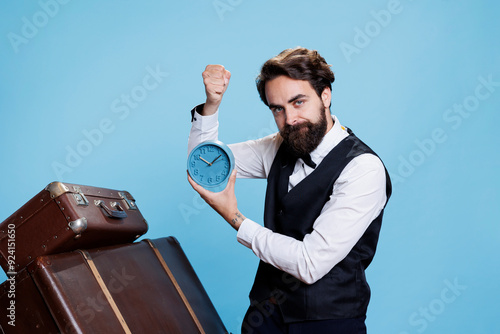 Hotel employee looks at time on camera, trying to be punctual in hospitality industry with wall clock. Professional doorkeeper in suit and tie appearing with watch against blue background. photo