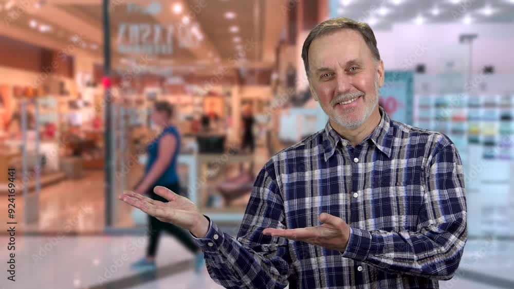 Smiling senior aged man standing indoors shows copy space. Shopping mall in the background. Smiling senior aged man standing indoors shows copy space. Shopping mall in the background.