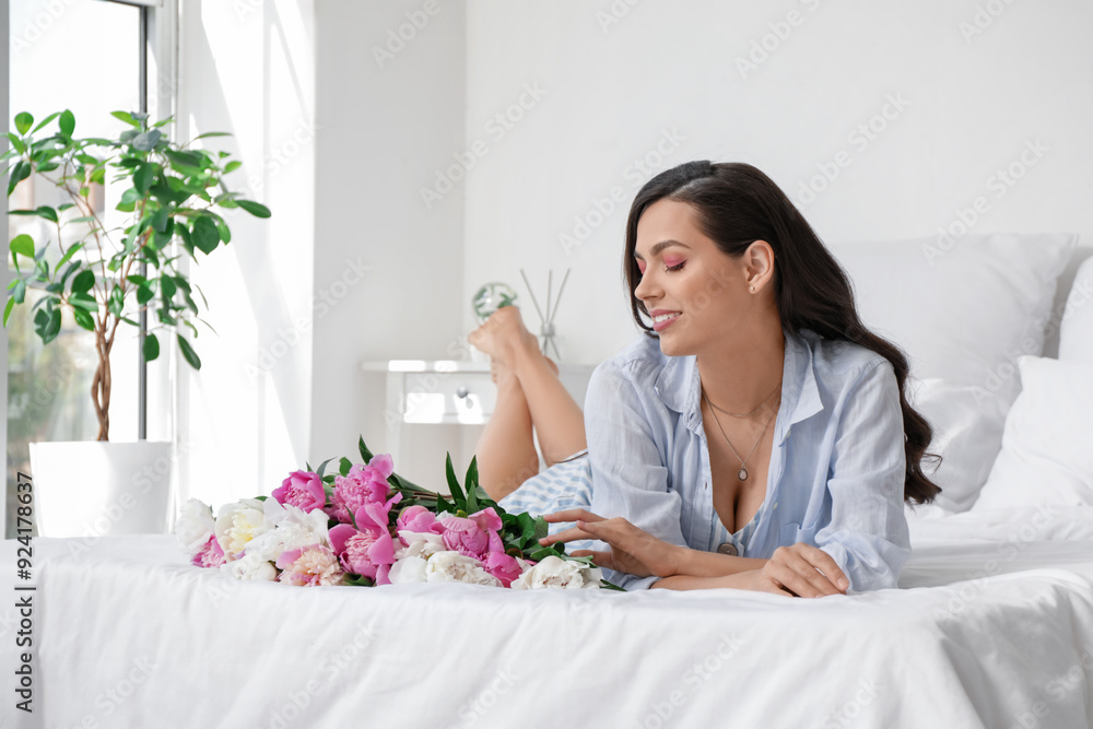 Happy young woman with bouquet of peony flowers lying on bed at home Happy young woman with bouquet of peony flowers lying on bed at home