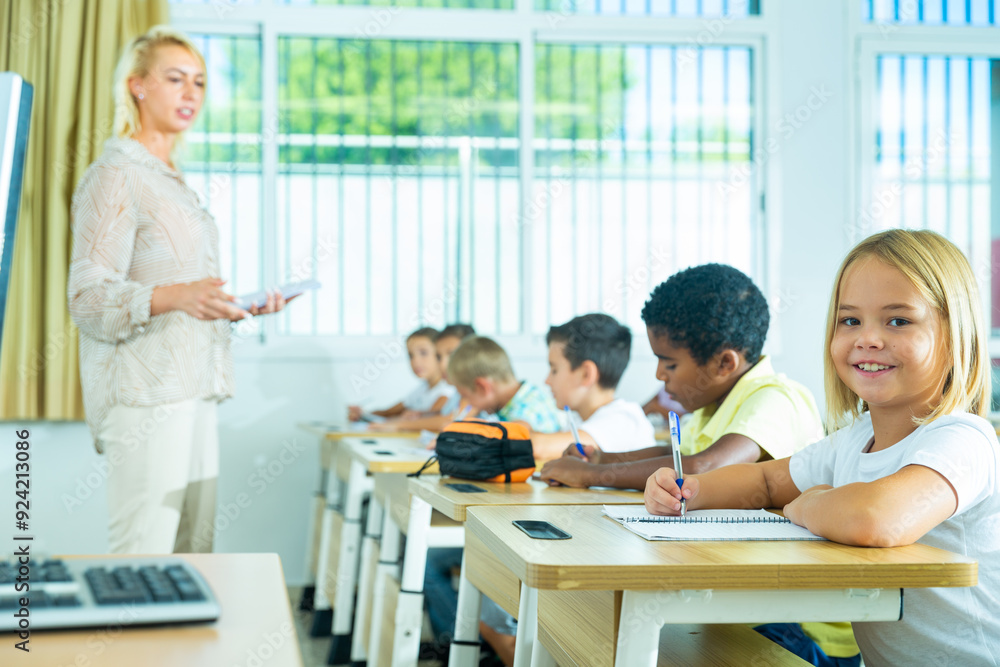Fototapeta premium Portrait of cheerful towheaded preteen schoolgirl sitting at desk in classroom during lesson,