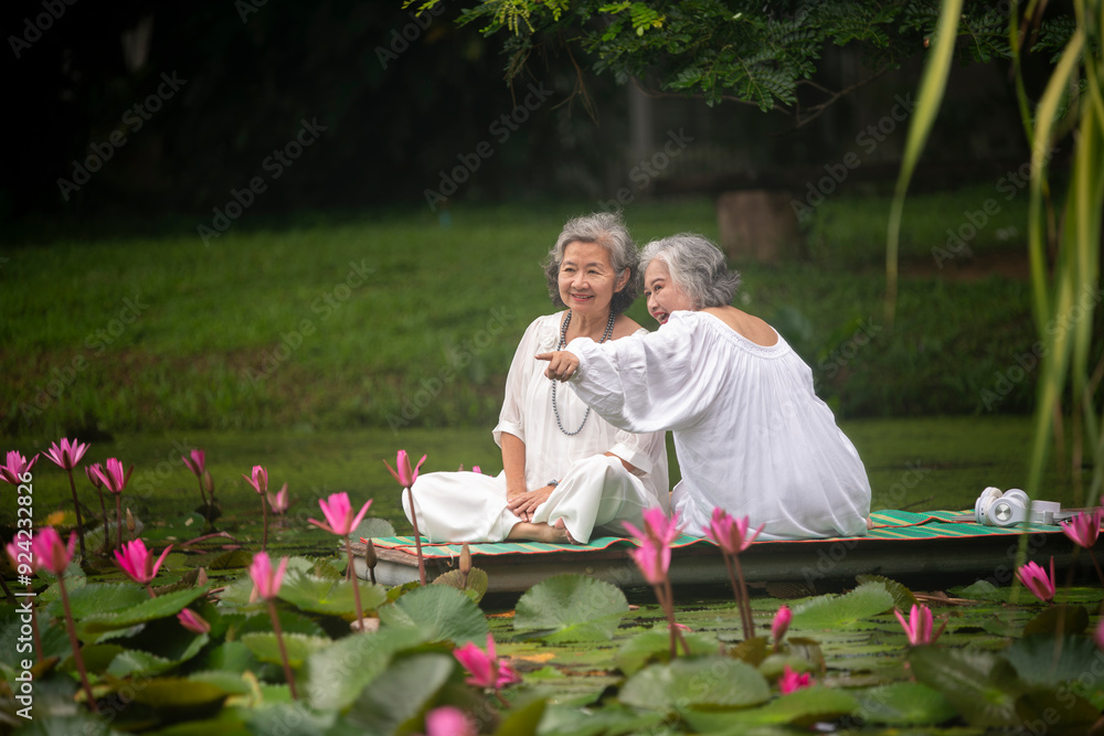 Naklejka premium two elderly women sitting by a pond surrounded by blooming lotus flowers. They are both wearing white tops and headphones, enjoying music while holding apples