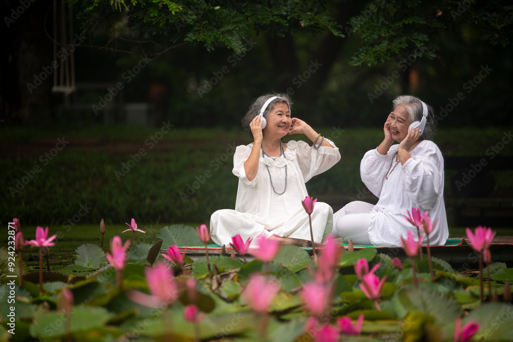 Naklejka premium two elderly women sitting by a pond surrounded by blooming lotus flowers. They are both wearing white tops and headphones, enjoying music while holding apples