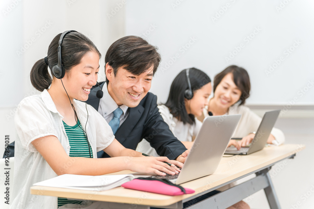 塾・学校の教室でパソコンとヘッドセットを使って先生に勉強を教わる小学生・中学生・高校生の子供
