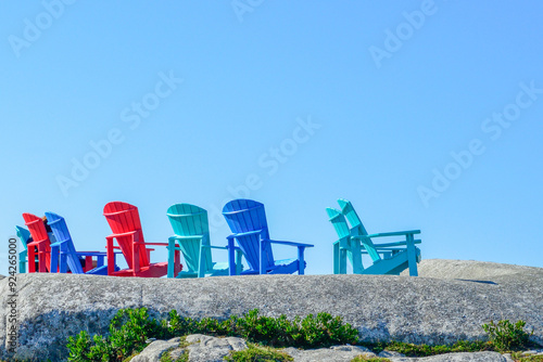 Multiple outdoor blue, red, and teal green polywood recycled plastic Adirondack chairs on a rocky cliff in a row overlooking the ocean. The sky is bright blue and the ground has patches of grass.  photo