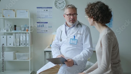 Middle aged doctor reading lab test results on clipboard and giving advice to young female patient, both sitting on examination table in medical office