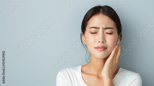 A young Asian woman with a pained expression, have a toothache and holding her cheek, with her eyes closed, standing against a light background.