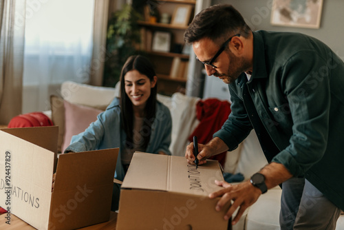 Focus man writing down on a cardboard box, preparing it for donations. Wife sorting things and sitting next to him