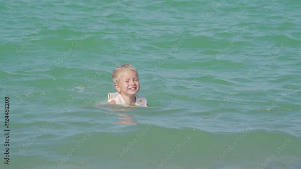 Happy little boy in swim vest life jacket playing on summer beach swimming in sea. Cute child in float lifejacket learning to swim in water. Yevpatoriya, Crimea, Russia 2024 Happy little boy in swim vest life jacket playing on summer beach swimming in sea. Cute child in float lifejacket learning to swim in water. Yevpatoriya, Crimea, Russia 2024