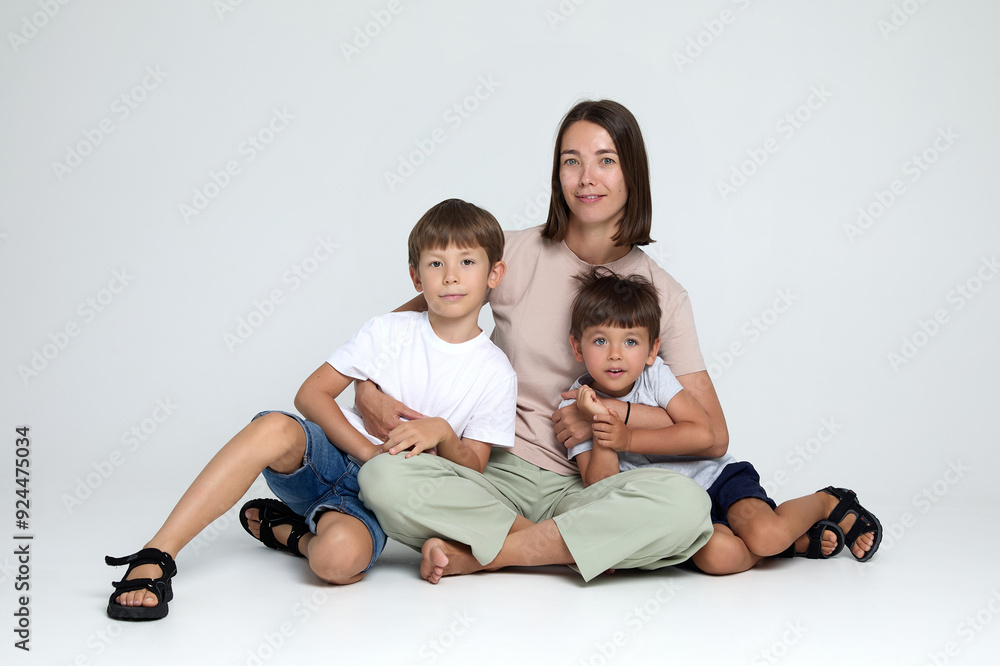 Full length photo of a smiling sons hugs his mother, they are sitting together, looking at camera, over grey background. Family concept. Full length photo of a smiling sons hugs his mother, they are sitting together, looking at camera, over grey background. Family concept.