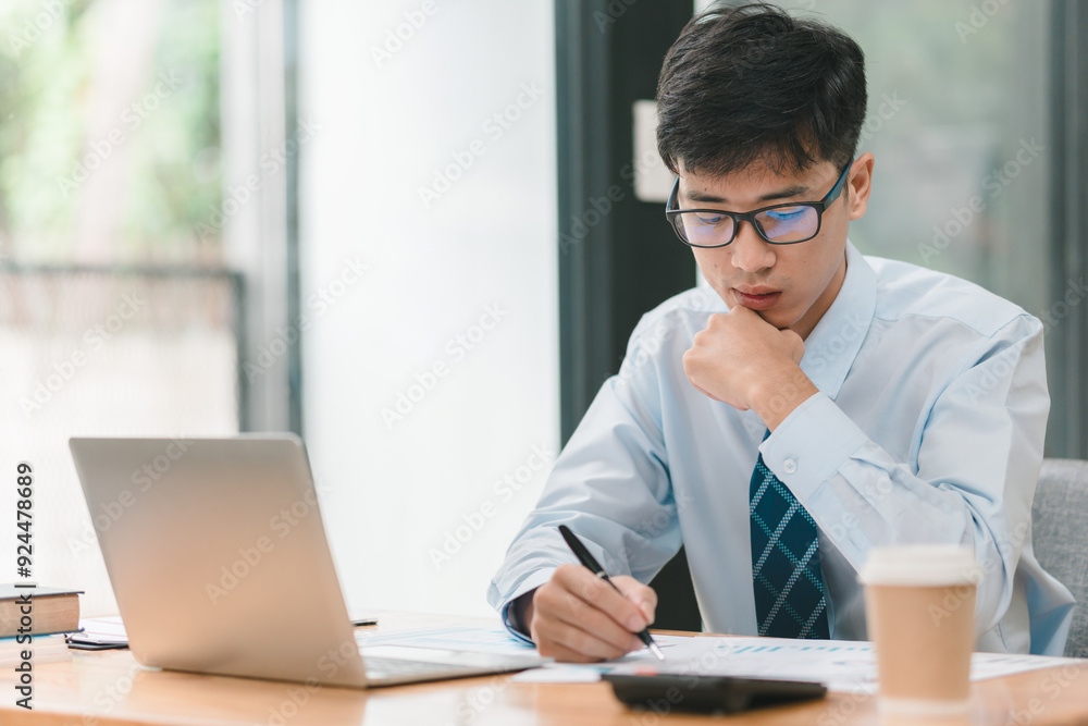 A man in a blue shirt and glasses is sitting at a desk with a laptop and a cup of coffee. He is writing on a piece of paper with a pen. The scene suggests a typical workday A man in a blue shirt and glasses is sitting at a desk with a laptop and a cup of coffee. He is writing on a piece of paper with a pen. The scene suggests a typical workday