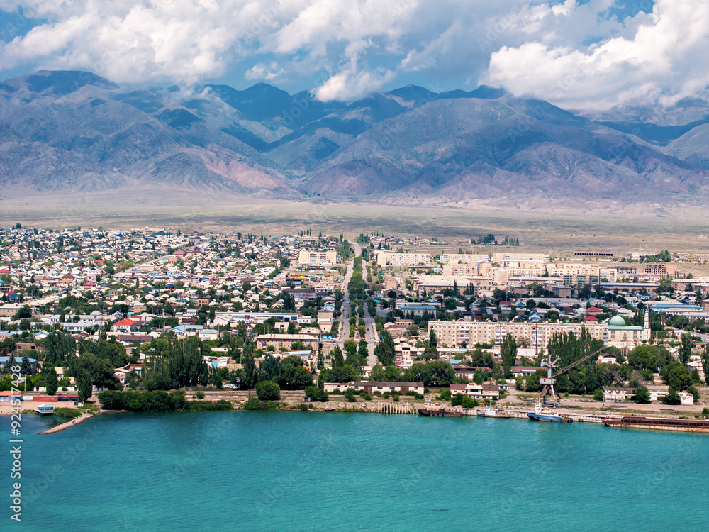 Naklejka premium Drone view on maritime city and port near mountains. View of the abandoned sea port with old crane, barge and tug in Central Asia. Vivid Issyk-Kul scenery.
