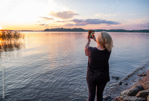 One caucasian woman on the lake shore admires the sunset and takes a selfie on a mobile phone.